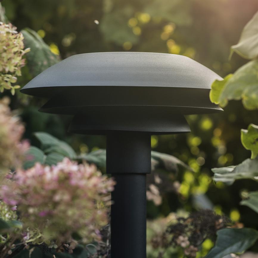 Eine schwarze, pilzartige Gartenbeleuchtungslampe steht auf einer dicken Säule, platziert zwischen grünen Blättern und rosa-weißen Blüten vor einem sonnenbeschienenen Gartenhintergrund.