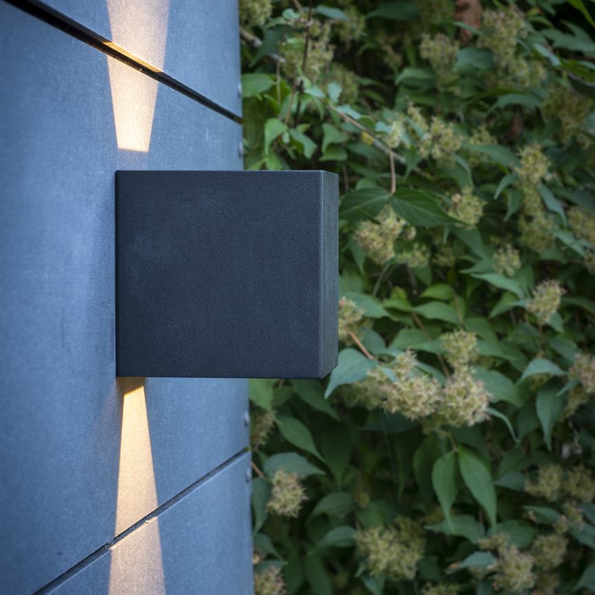 Eine schwarze, quadratische Wandleuchte strahlt warme Lichtstrahlen nach oben und unten aus, montiert an einer grau gefliesten Wand neben einer üppig grünen Hecke mit leuchtenden Blumen.
