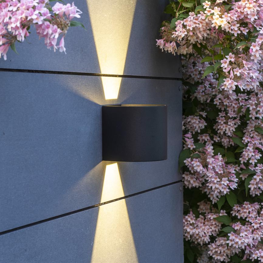 Sort cylindrisk væglampe udsender smalle, koniske lysstråler opad og nedad på en grå panelvæg ved siden af en tæt hæk med små lyserøde blomster.