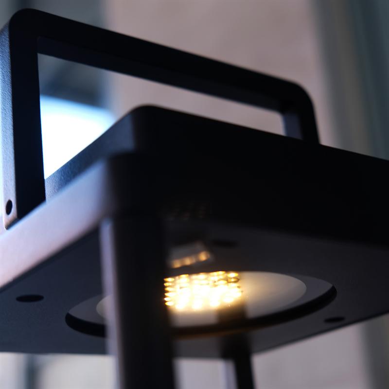 Schwarze, quadratische Laterne mit Griff strahlt warmes, gelbes LED-Licht aus; Es ist auf einem Metallrahmen in einer verschwommenen Innenumgebung vor einem Fenster montiert.