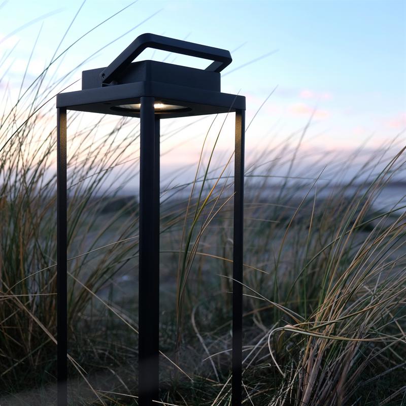Eine schwarze Metalllaterne steht im Sand und wirft in der Abenddämmerung schwaches Licht nach unten zwischen hohen Strandgräsern, mit einem sanften pastellfarbenen Himmel und Meer im Hintergrund.