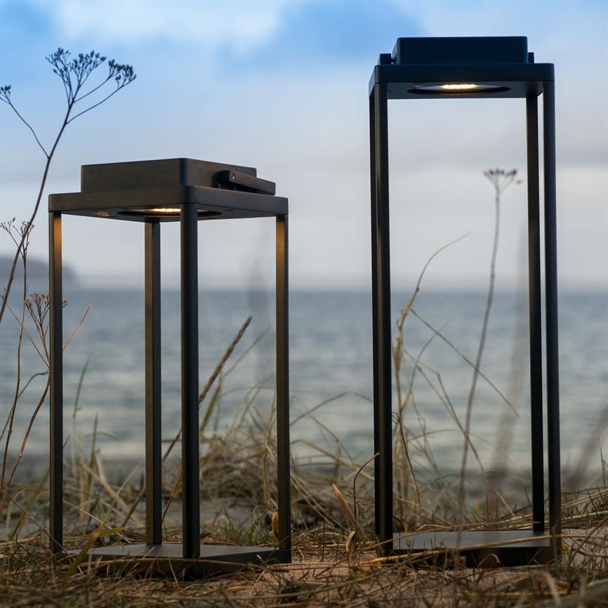 To sorte, rektangulære metallanterner står svagt tændt på en sandklit med tørre strå og vilde blomster, foran et roligt hav under en lys, skyet himmel.
