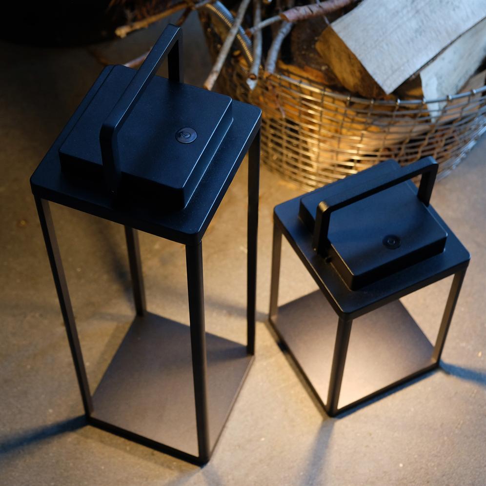 Zwei schwarze Metalllampen stehen nebeneinander auf dem Betonboden und werfen sanfte Schatten; Im Hintergrund sieht man einen Weidenkorb mit Brennholz und einigen Zweigen.