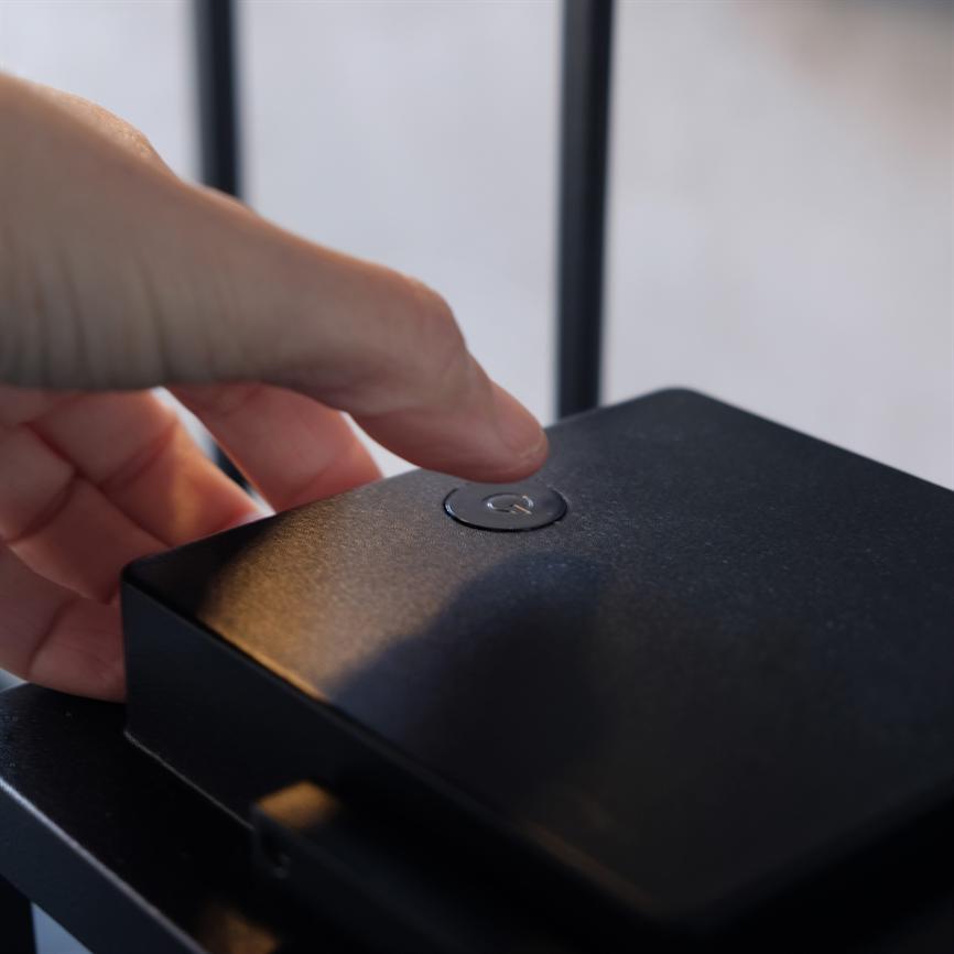 En finger trykker på en rund tænd/sluk-knap på en lille sort elektronisk boks, placeret på en hylde med sløret, lyst baggrund og to lodrette antennestænger.