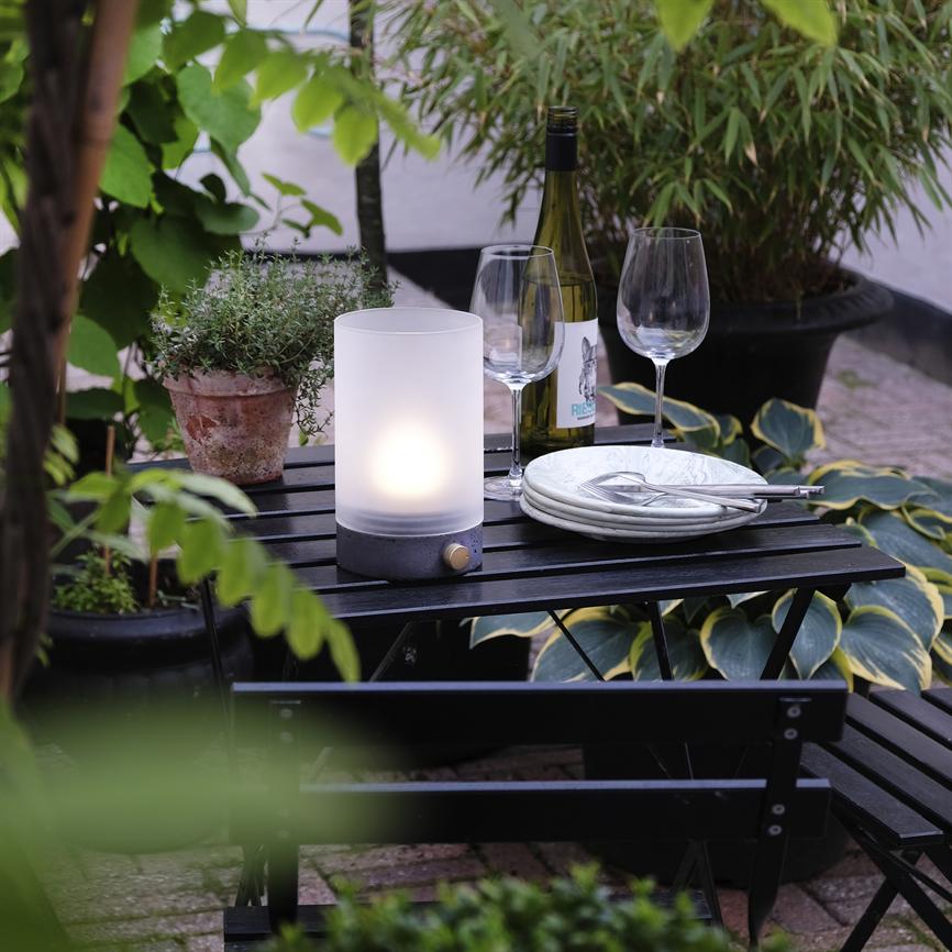 Eine weiße Tischlampe leuchtet auf einem kleinen schwarzen Cafétisch, umgeben von zwei Weingläsern, einer Weinflasche, einem Stapel Tellern und Topfpflanzen auf einer Außenterrasse.