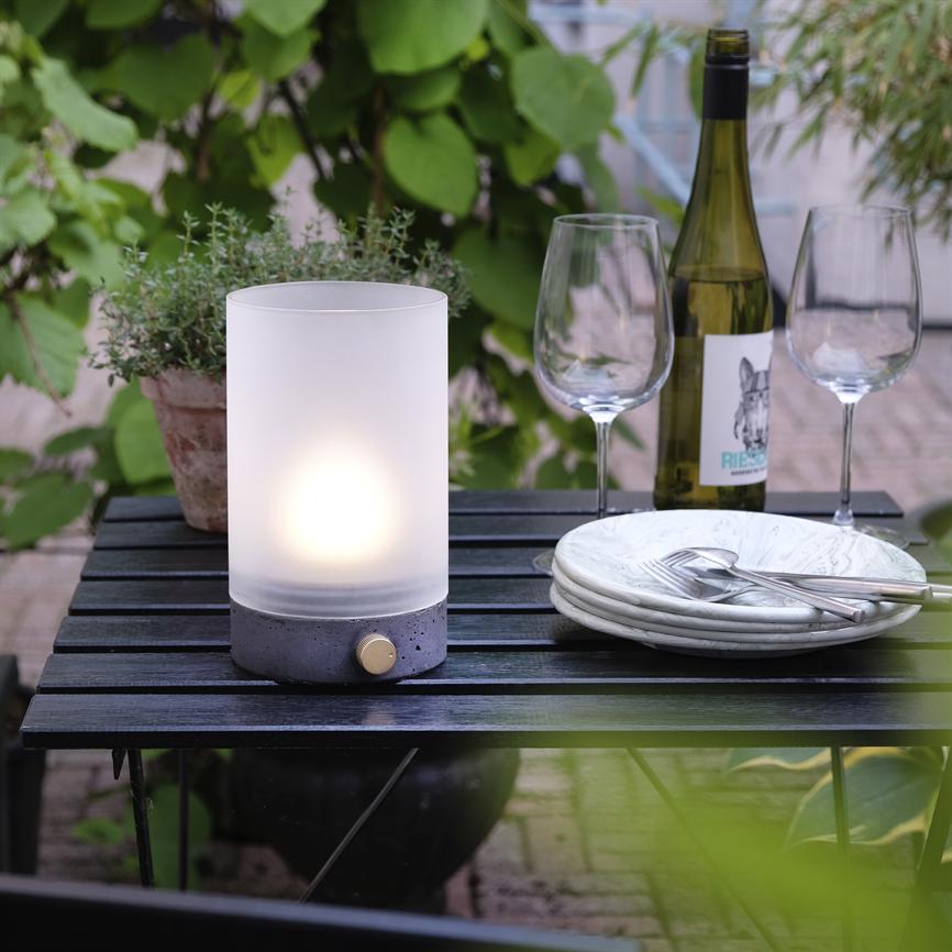Zylindrische Tischleuchte strahlt sanftes Licht aus; steht auf einem schwarzen Holztisch neben zwei Weingläsern, einer Weinflasche und einem Stapel Teller mit Besteck, umgeben von grünen Pflanzen. Text auf dem Etikett: „RIES“.