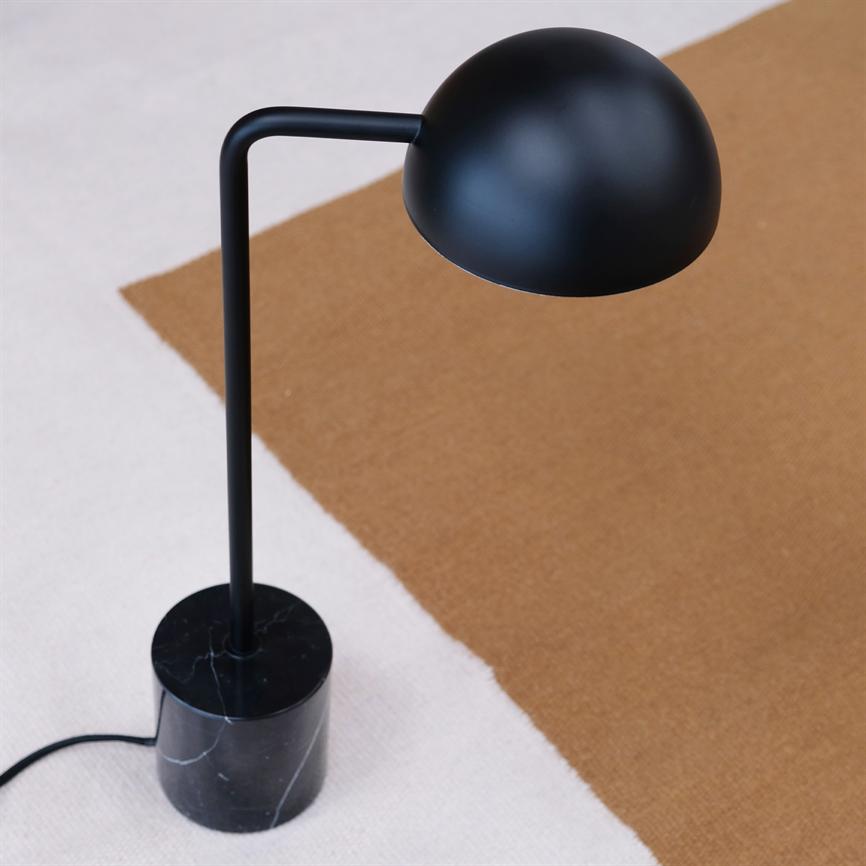 Sort bordlampe med rund skærm og sort marmorfod står på et lystgråt og brunt tæppe; ledning ligger delvist synlig langs kanten.