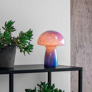 Die pilzförmige Glaslampe strahlt ein warmes, spiralförmig gestreiftes Licht aus und steht auf einem schwarzen Metallregal zwischen Sukkulenten in Töpfen vor einer hellen Wand mit einer Holzplatte auf der rechten Seite.