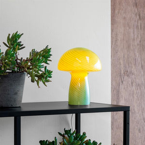 Eine pilzförmige Glaslampe in Gelbgrün leuchtet sanft auf einem schwarzen Metallregal, umgeben von Sukkulenten in grauen Töpfen vor einer weißen Wand mit einer Holzplatte rechts davon.