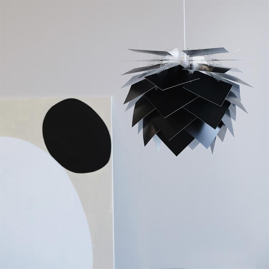 Von der Decke hängt eine schwarz-silberne Pendelleuchte; Seine überlappenden Metallplatten bilden eine kegelartige Form. In einem hellen, minimalistischen Raum neben einem abstrakten Gemälde mit einem großen schwarzen Kreis.