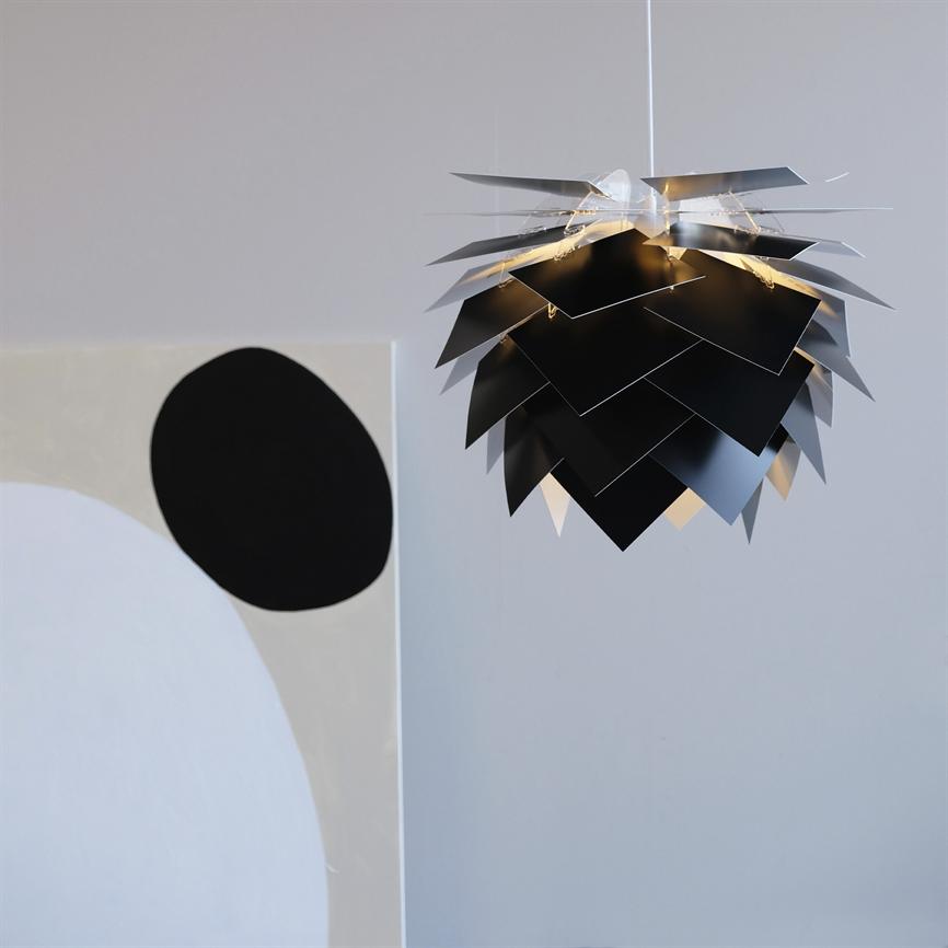 Sort, skællignende pendelarmatur hænger i loftet og kaster varmt lys mellem overlappende plader; omgivet af en lys, minimalistisk væg med abstrakt maleri med sorte og hvide former.