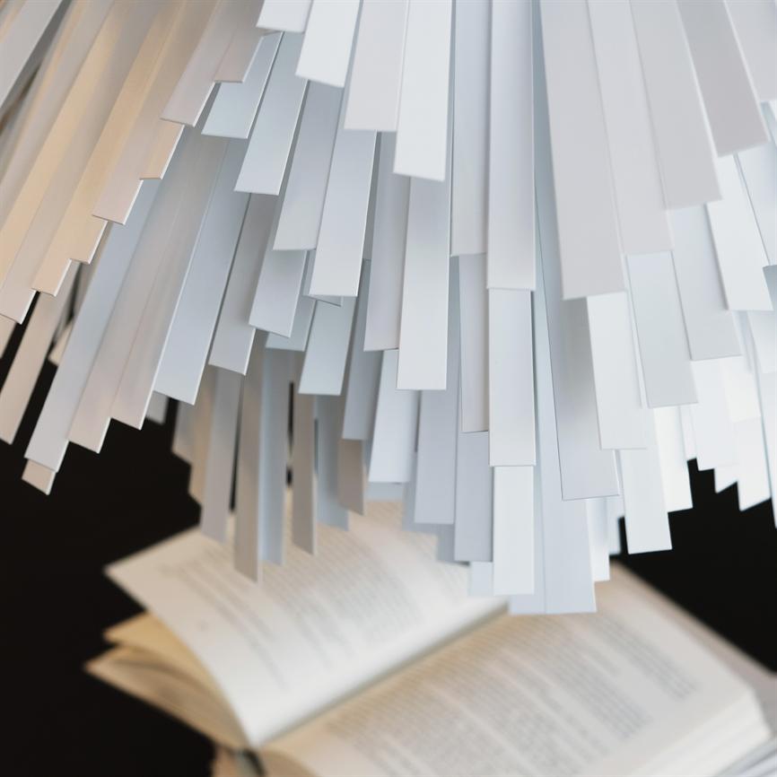 Weiße Skulptur/Lampe aus hängenden rechteckigen Papierstreifen; Es hängt über einem aufgeschlagenen Buch mit undeutlichem Text auf dunklem Hintergrund.