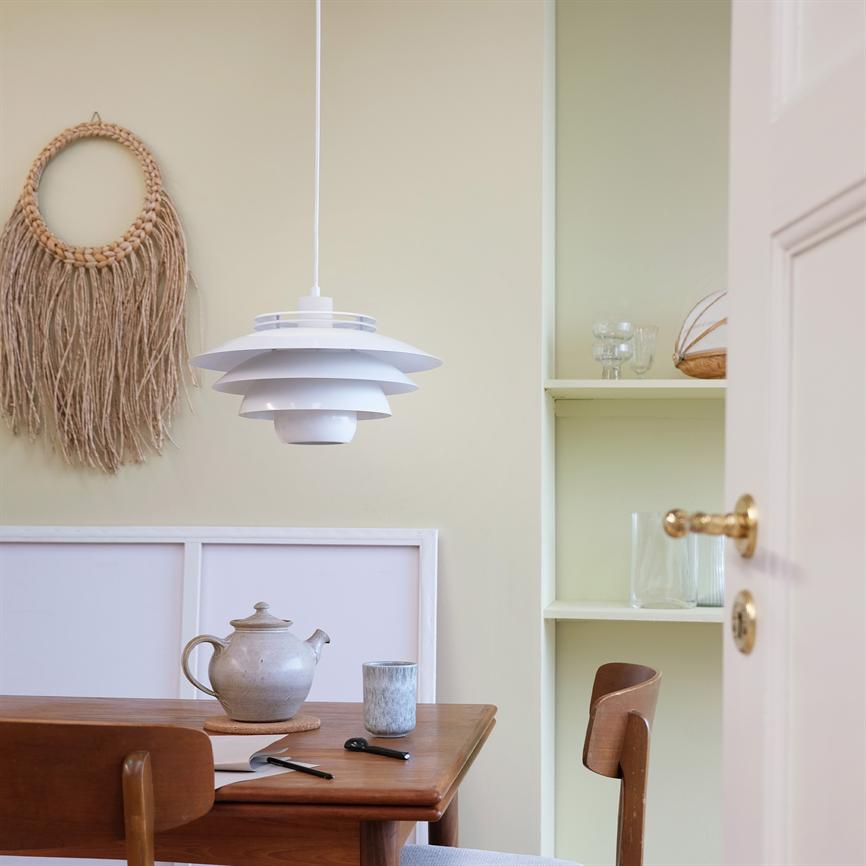 Eine weiße Pendelleuchte hängt über einem Holztisch, auf dem eine Teekanne und eine Tasse aus Keramik stehen. umgeben von einer Makramee-Wanddekoration, offenen Regalen mit Glas und einer teilweise geöffneten Tür.