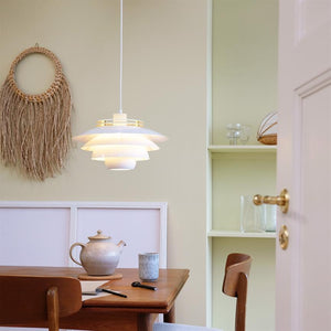 Eine weiße, mehrschichtige Hängelampe beleuchtet einen Holztisch in einer hellen Essecke mit Teekanne, Becher, zwei Holzstühlen, Makramee-Wanddekoration und eingebauten Regalen.