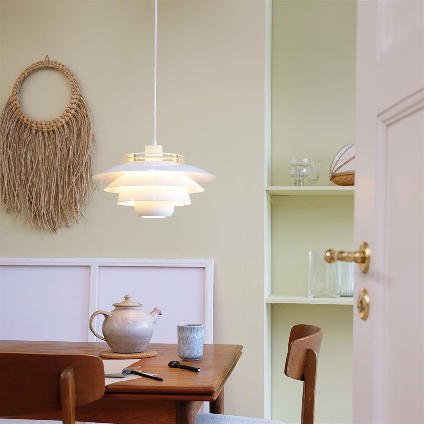 Eine weiße, mehrschichtige Hängelampe beleuchtet einen Holztisch in einer hellen Essecke mit Teekanne, Becher, zwei Holzstühlen, Makramee-Wanddekoration und eingebauten Regalen.