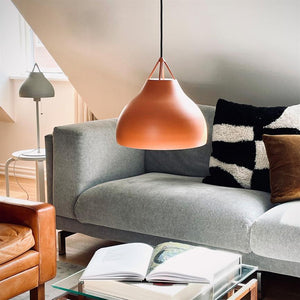 Eine orangefarbene Pendelleuchte hängt über einem aufgeschlagenen Buch auf einem Couchtisch aus Glas; In einer hellen, modernen Wohnzimmerecke steht ein graues Sofa mit einem schwarz-weiß gemusterten Kissen und einem braunen Samtkissen.