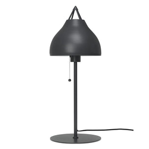 Tischleuchte mit dunkelgrauem, abgerundetem Schirm hängt über einer vertikalen Stange und wird per Zugknopf bedient; auf rundem Sockel mit sichtbarer Kordel platziert, vor neutralweißem Hintergrund.
