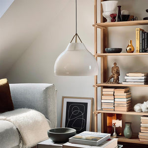 Über einem kleinen Couchtisch hängt eine weiße Glaspendelleuchte; im Hintergrund ein hellgraues Sofa mit Plaid und ein hölzernes Bücherregal voller Bücher, Keramik und einer kleinen Skulptur.