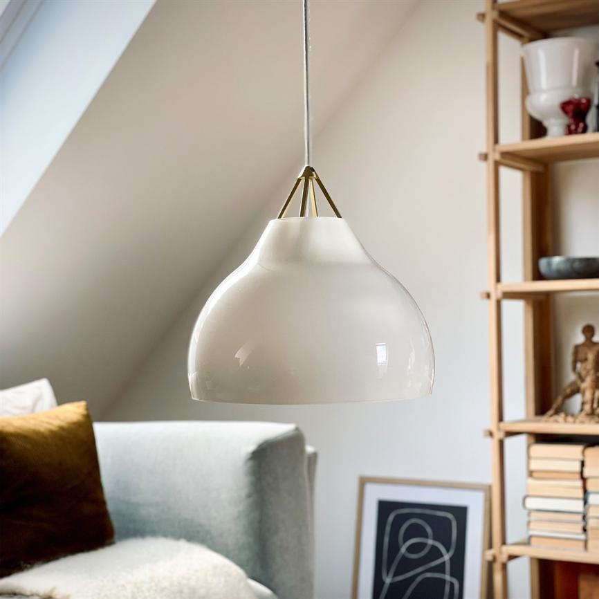 En hvid glaspendel hænger roligt i snor; den lyser ikke og reflekterer dagslys. Baggrund: lyse skrå vægge, lysegrå sofa med gylden pude, reol med bøger og dekorative genstande.