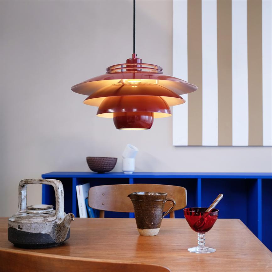 Eine rote mehrschichtige Pendelleuchte hängt und wirft warmes Licht über einen Holztisch mit Teekanne, Keramikbecher und roter Glasschale; hinter einem blauen Bücherregal und vertikalen beige-weißen Streifen an der Wand.