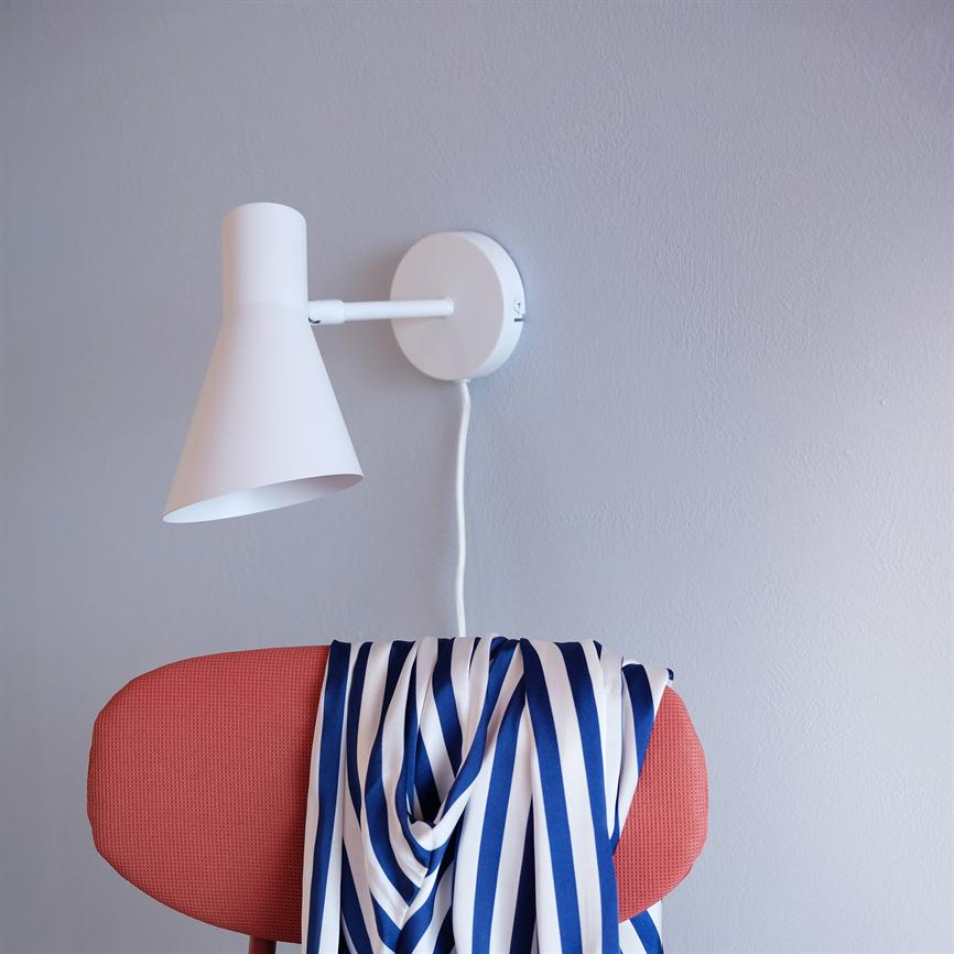 Weiße Wandleuchte, montiert an einer hellgrauen Wand mit sichtbarem weißem Kabel; Über der Rückenlehne eines rot gepolsterten Stuhls hängt ein blau-weiß gestreiftes Stück Stoff.
