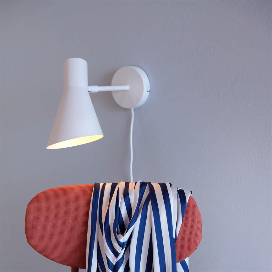 Eine Wandlampe scheint auf einen rosa gepolsterten Stuhl, über dessen Rückenlehne lässig ein blau-weiß gestreifter Schal hängt, vor einer hellgrauen Wand.