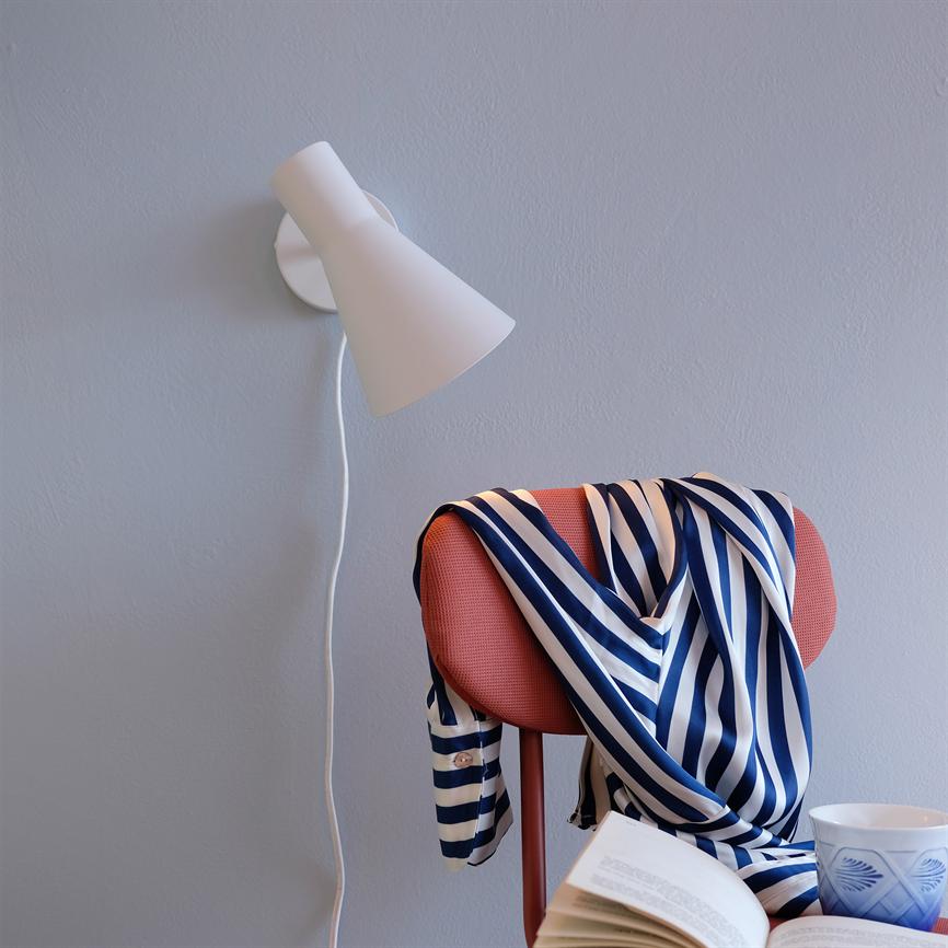 Hemd mit blau-weißen Streifen hängt über einer korallenroten Rückenlehne; Eine weiße Wandlampe scheint herab, ein aufgeschlagenes Buch und eine Tasse stehen auf der Sitzbank in einem hellgrauen Raum.