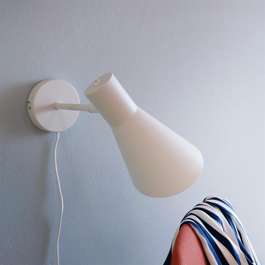 Weiße Wandleuchte zeigt nach unten auf einen korallenfarbenen Stuhl; Über der Stuhllehne hängt ein blau-weiß gestreifter Schal, im Hintergrund eine hellgraue Wand und sichtbarer Draht.