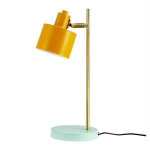 Gelbe Tischlampe mit schrägem Schirm, montiert auf einem schlanken Messingstab und mintgrünem Rundfuß; Schwarzes Kabel verläuft seitlich auf weißem Hintergrund.