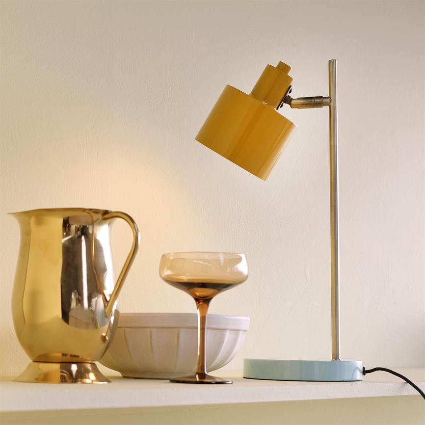 Eine nach unten geneigte gelbe Tischlampe wirft sanftes, warmes Licht auf einen goldenen Krug, ein bernsteinfarbenes Cocktailglas und eine weiße Keramikschale auf einem hellen Regal vor einer cremefarbenen Wand.
