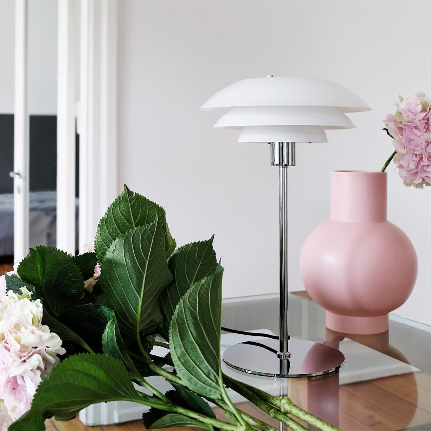 Weiße, mehrschichtige Tischleuchte steht auf einem Glastisch neben einer rosa Vase; Grüne Hortensienblätter und rosa Blüten sind verstreut, Hintergrund mit weißer Wand und offenem Raum.