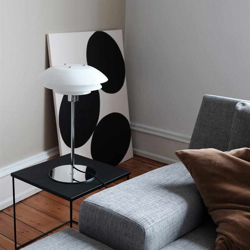 Ausgeschaltete Tischlampe auf schwarzem Beistelltisch; daneben lehnt ein weißes Gemälde mit drei großen schwarzen Kreisen an der Wand, vor einem grauen Sofa auf einem Holzboden.