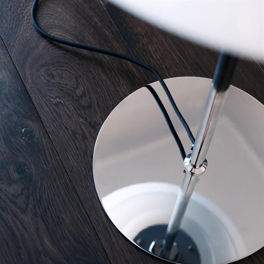 Chromfuß einer Stehlampe steht auf dunklem Holzboden; Ein glänzender, runder Sockel spiegelt den Lampenstiel wider, während sich eine schwarze, geflochtene Kordel über den Sockel und den Boden schlängelt.