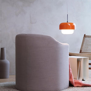 Ein rosafarbener Polstersessel ist abgewandt, über einem niedrigen Beistelltisch mit Büchern leuchtet eine orangefarbene Hängelampe; Ein Korbstuhl, ein Teppich und eine Vase stehen in einem ruhigen, minimalistischen Raum.