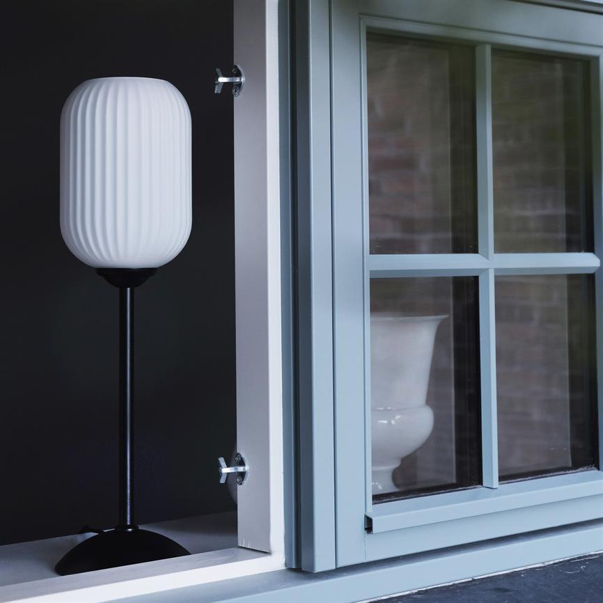 Eine weiße gerippte Lampe steht auf einem schwarzen Fuß, platziert neben einem offenen hellblauen Fenster mit Gittern; Dunkler Hintergrund und eine weiße Vase, die durch das Fenster gesehen wird.