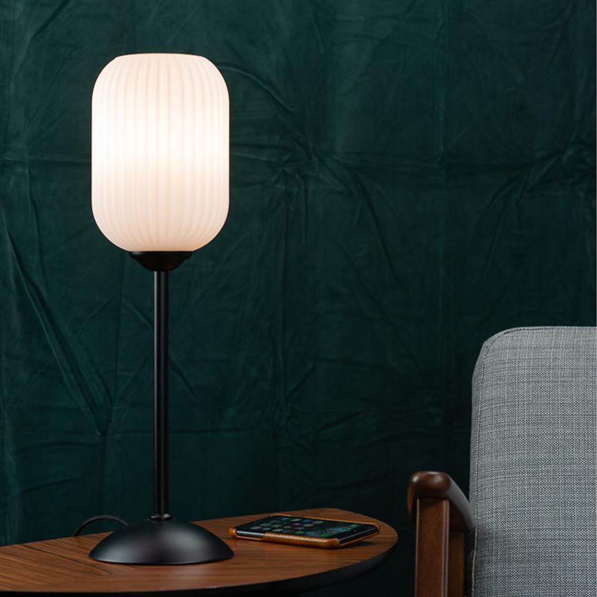 Sanft leuchtende Tischlampe auf kleinem Holztisch mit Smartphone; neben einem grauen Polsterstuhl vor einem dunkelgrünen Strukturhintergrund.