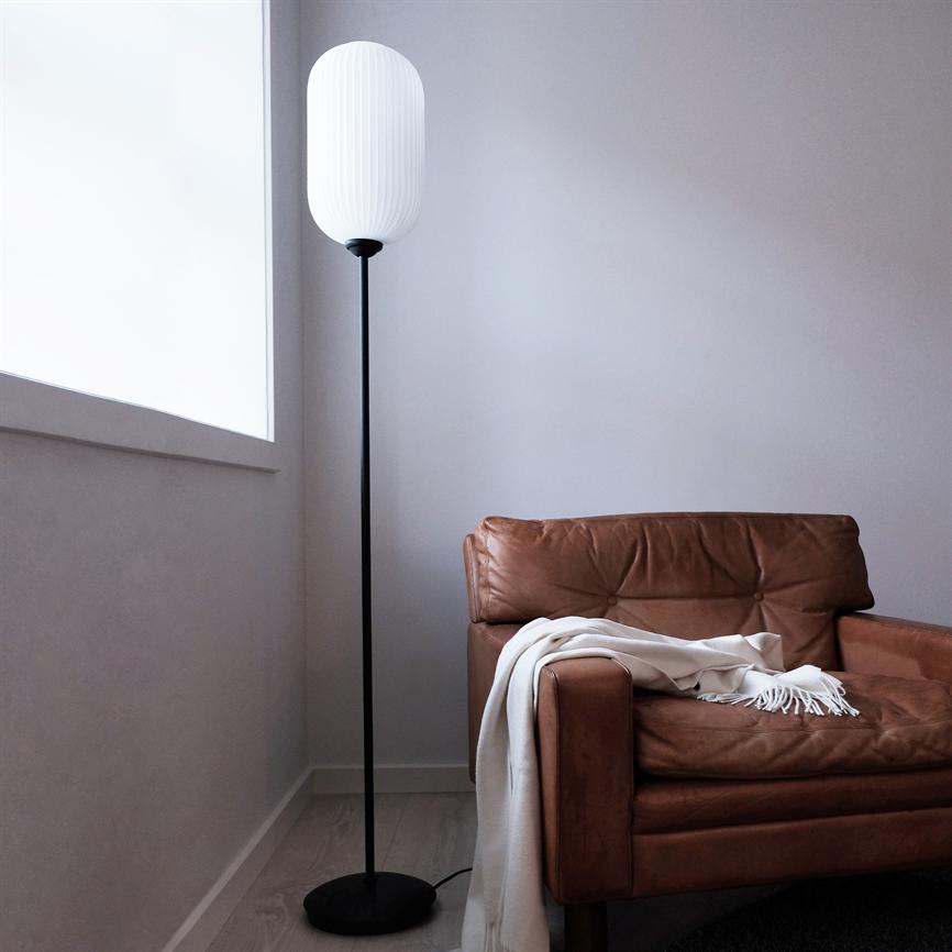 Brauner Ledersessel steht in einer Ecke, über der Armlehne liegt ein weißer Teppich; Daneben steht eine hohe Stehlampe mit weißem Schirm, umgeben von einem hellen Holzboden, grauen Wänden und einem großen Fenster.