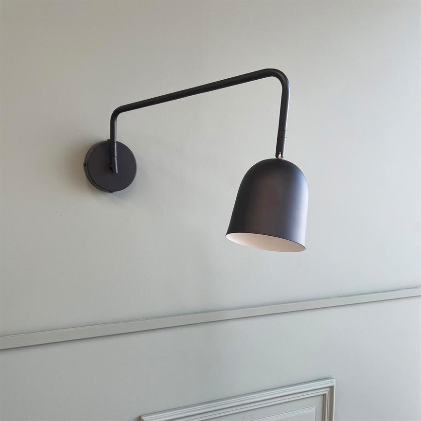 Eine schwarze Wandleuchte hängt an einem langen, gebogenen Arm und ist nach unten geneigt, mit weißer Innenseite; Es wird an einer hellgrauen Wand über einer dezenten Paneelleiste montiert.