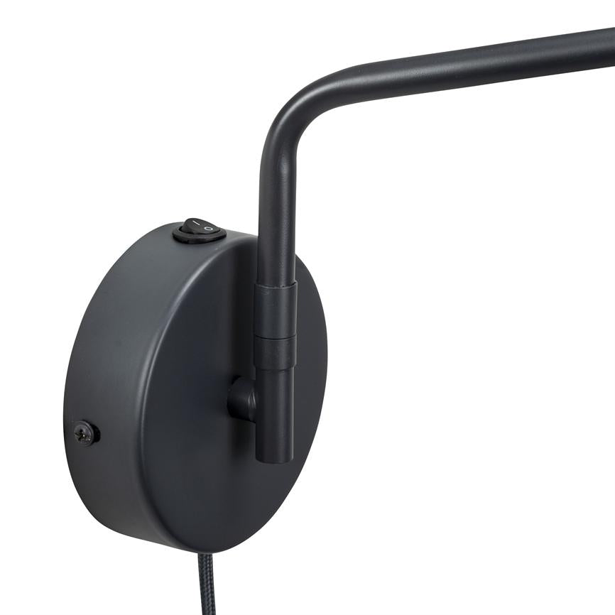 Die schwarze, runde Wandhalterung für die Lampe hält einen gebogenen, verstellbaren Arm; Oben sitzt ein kleiner Umschaltknopf mit der Aufschrift „O“, nach unten führt ein mit Stoff ummanteltes Kabel vor weißem Hintergrund.