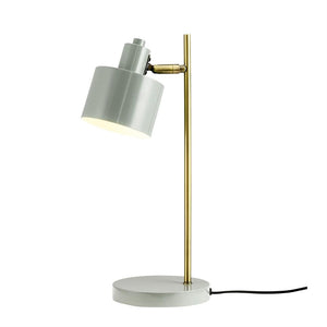 Eine Tischlampe mit grauem zylindrischem Schirm und Messingstab; Der Bildschirm zeigt schräg nach unten und strahlt weiches Licht aus, platziert auf einem weißen Hintergrund.