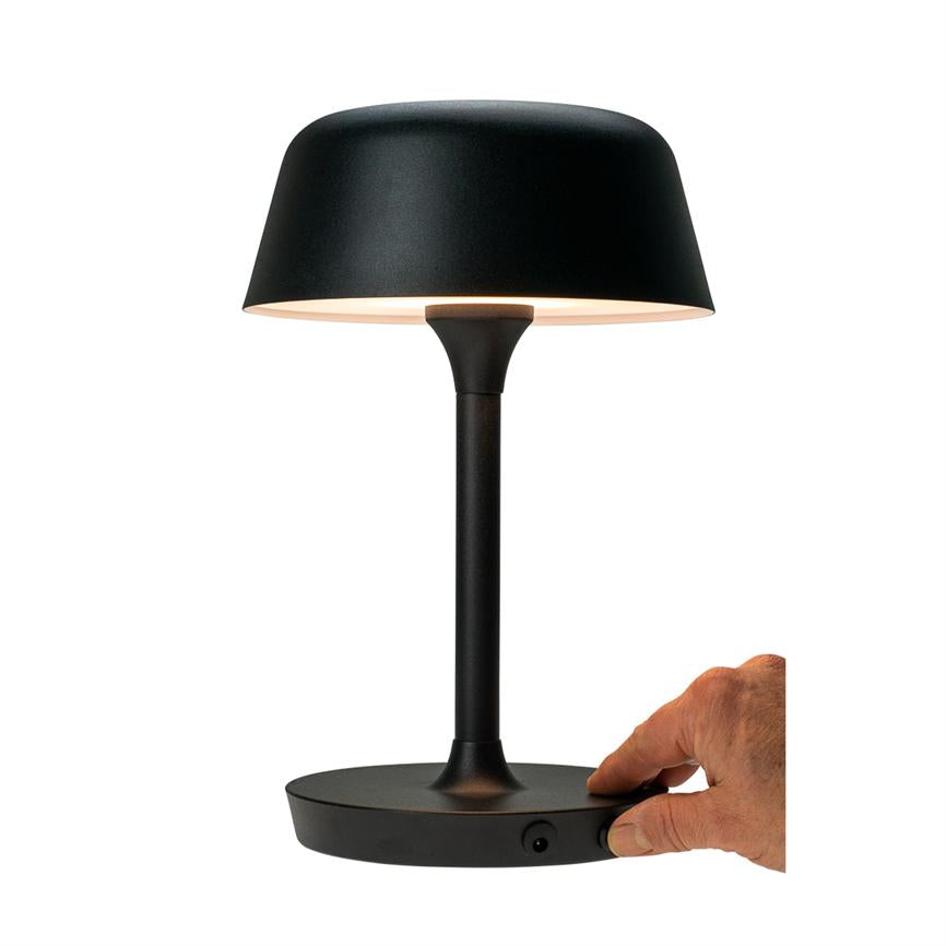 Schwarze Tischleuchte mit rundem, nach unten leuchtendem Lampenschirm; eine Hand dreht einen Knopf am flachen Sockel der Lampe; Die Lampe steht auf einer ebenen Fläche vor einem neutralweißen Hintergrund.