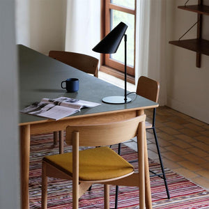 Auf einem grünen Esstisch steht eine schwarze Tischlampe, dazu eine aufgeschlagene Zeitschrift und eine blaue Tasse; umgeben von Holzstühlen, einem gestreiften Teppich und einem Fenster, das natürliches Tageslicht hereinlässt.