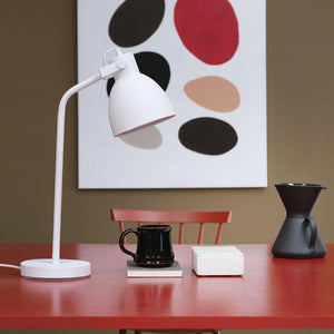 Weiße Schreibtischlampe zeigt auf eine schwarze Tasse auf einer roten Tischplatte, flankiert von einer weißen Schachtel und einem schwarzen Kaffeetrichter; roter Stuhl und abstrakte Malerei im Hintergrund.