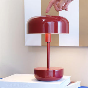 Rote Tischlampe mit rundem Schirm; eine Hand greift nach unten und drückt den Knopf oben; Die Lampe steht auf einem Stapel weißer Bücher vor einer hellen Wand mit einem Gemälde. 
 Text zum Buch: „IllustrativeBranding™“