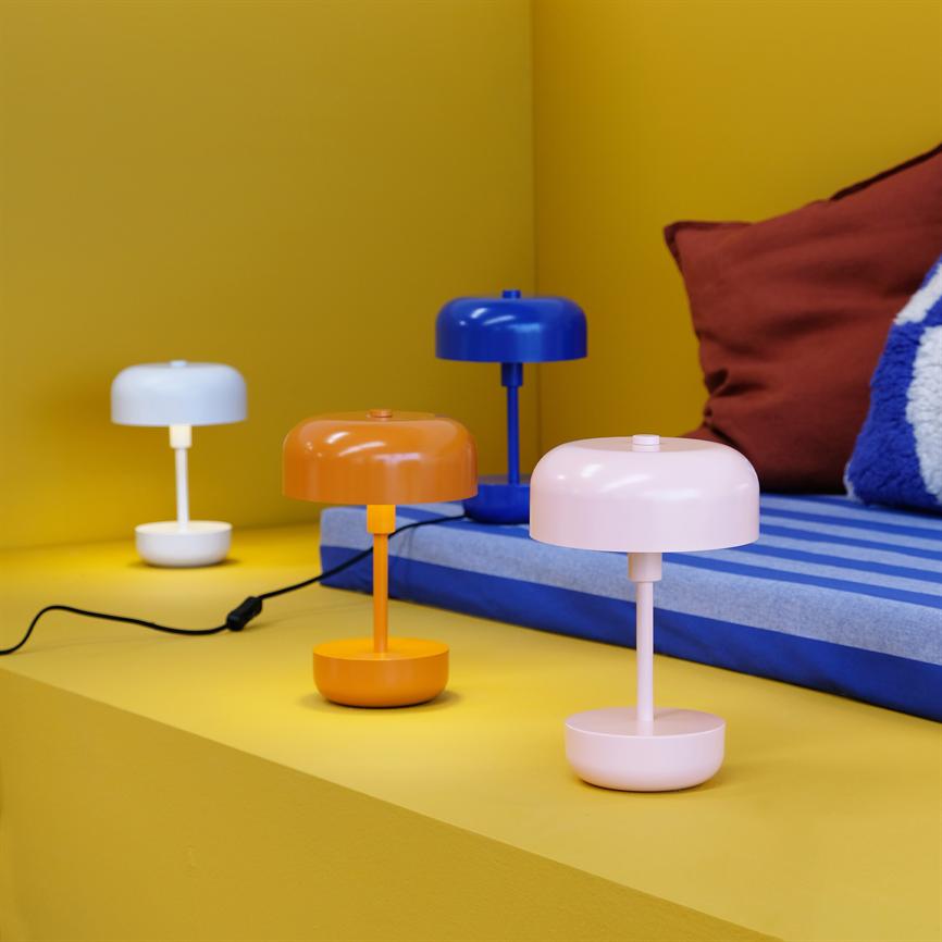 Vier kleine, moderne Tischlampen in Weiß, Orange, Blau und Pink stehen auf einem gelben Regal vor einer blau gestreiften Matratze und einem braunen Kissen.