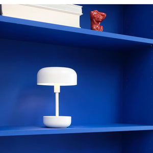 Weiße, moderne Tischlampe leuchtet schwach; steht auf einem leuchtend blauen Bücherregal mit einem Stapel weißer Bücher und einer kleinen roten Keramikfigur auf dem Regal darüber.