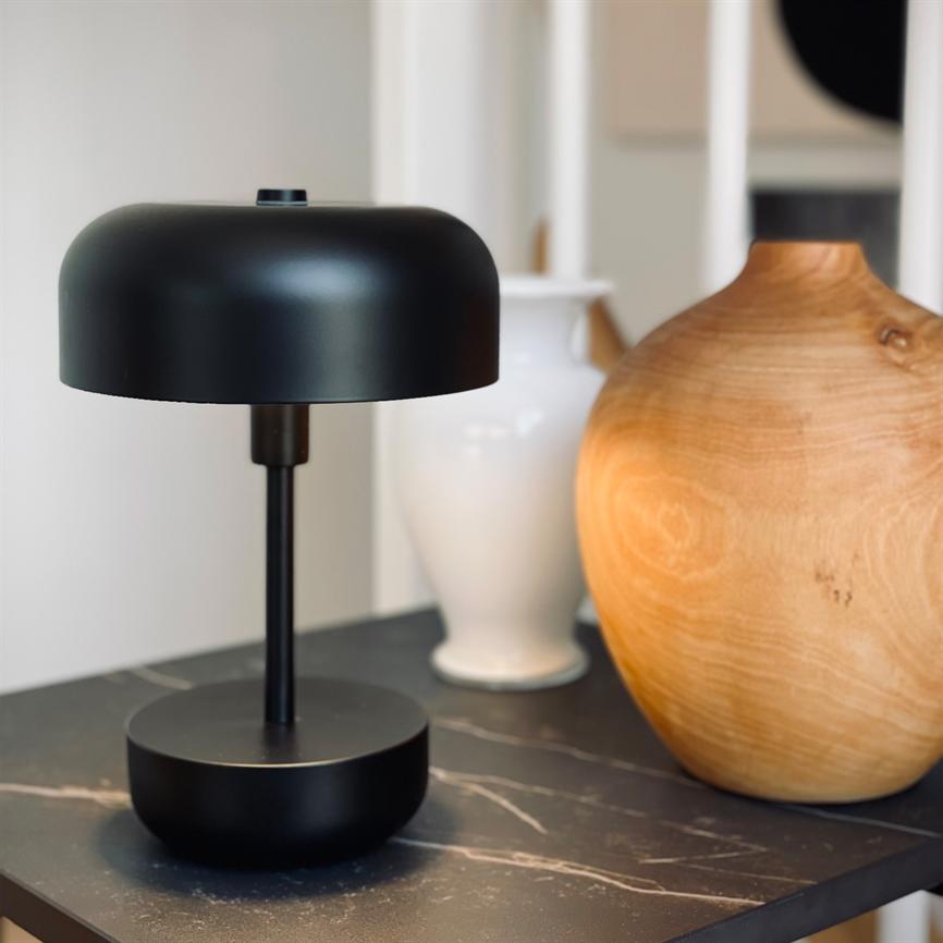Auf einem dunklen Marmortisch steht eine schwarze Tischlampe, flankiert von einer weißen Vase und einer runden Holzvase in einer hellen, modernen Ecke des Wohnzimmers.