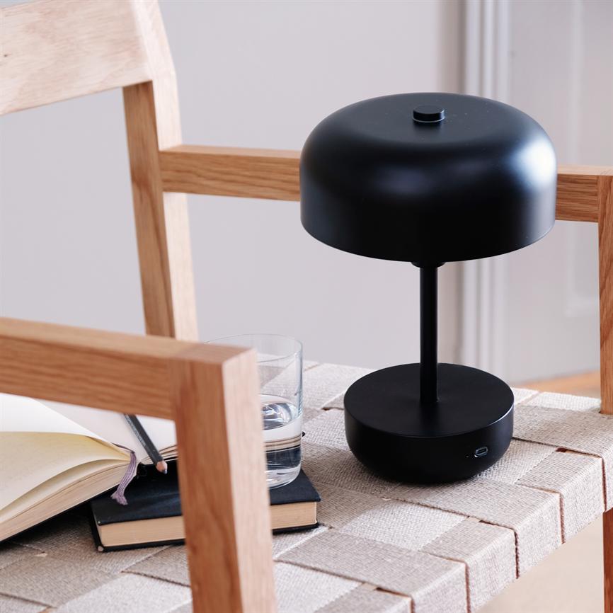 Schwarze, moderne Tischlampe steht auf der Korbsitzfläche eines Holzstuhls; Daneben liegen Notizbücher, ein Bleistift und ein Glas Wasser in einem hellen, minimalistischen Raum.