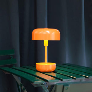 Eine orangefarbene Tischlampe mit rundem Schirm leuchtet sanft und steht auf einem grün lackierten Holztisch vor dunklen Vorhängen.