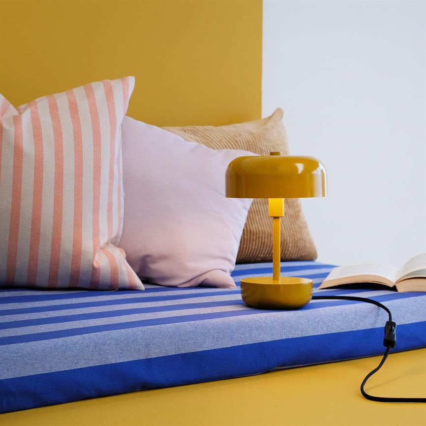 Eine gelbe Tischlampe steht auf einem blau-hellblau gestreiften Kissen neben einem aufgeschlagenen Buch; drumherum liegen rosa, gestreifte und beige Kissen vor gelb-weißem Hintergrund.